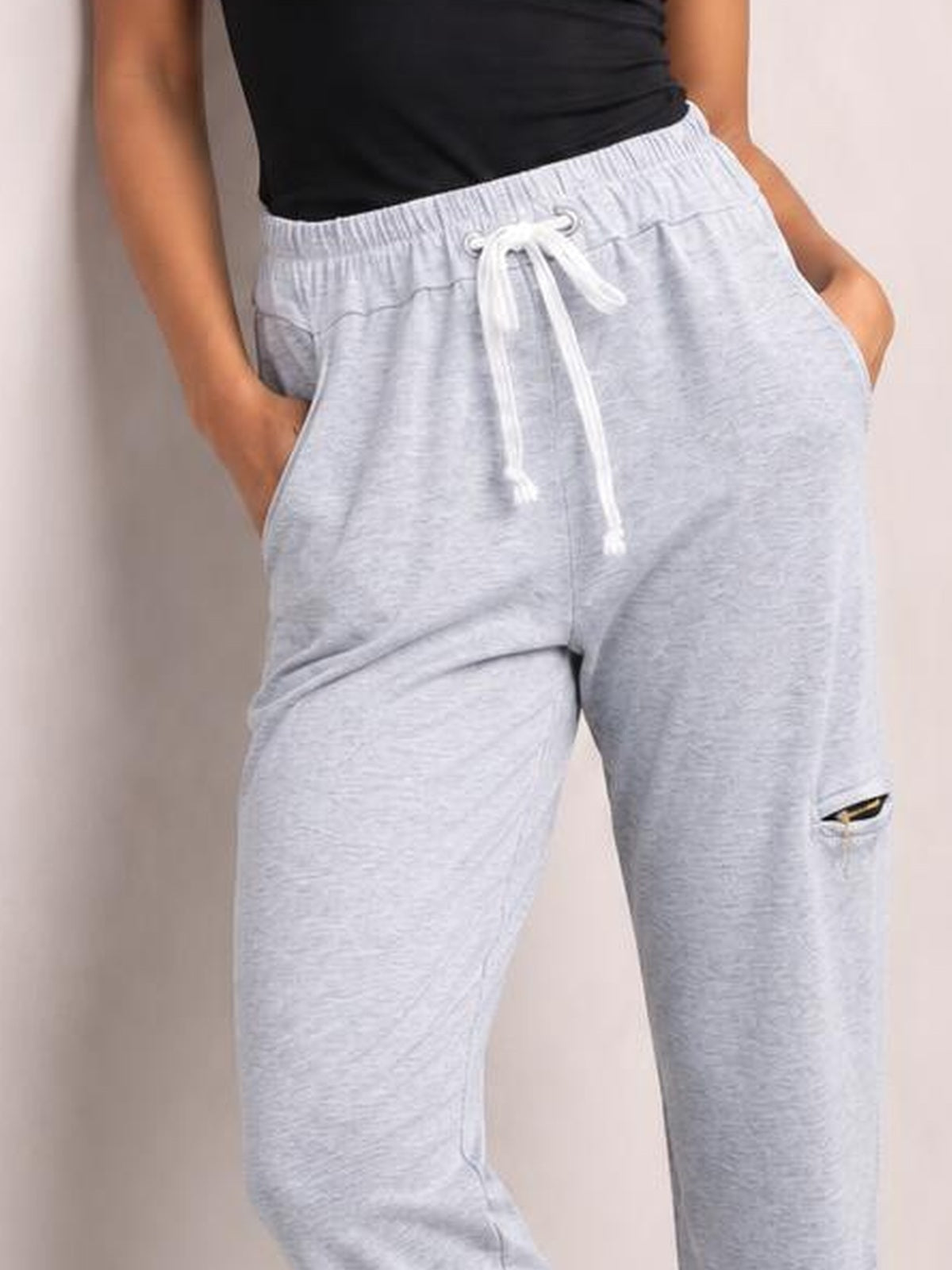 A model wears light gray drawstring joggers with an elastic waistband, relaxed straight-leg silhouette, side pockets, and a visible zip pocket detail.
