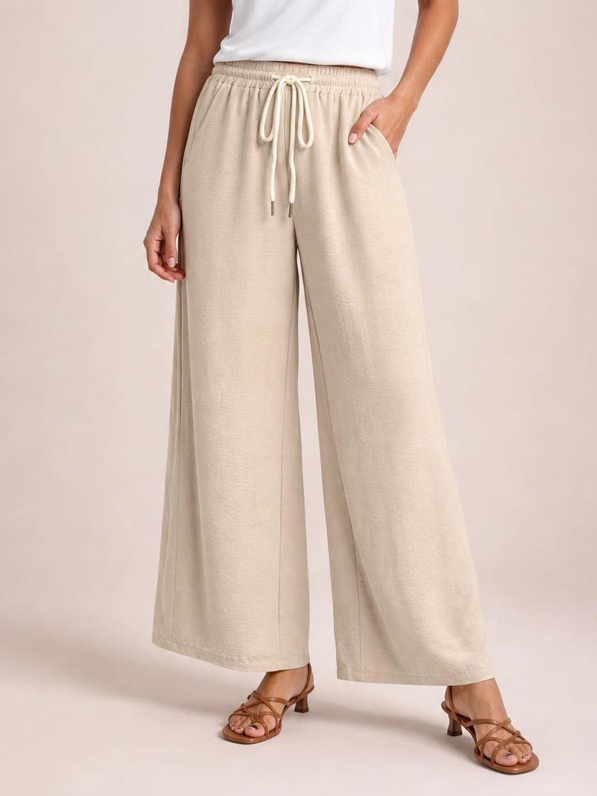 A model stands in beige drawstring wide leg pants with an elastic waist, front pockets, and a relaxed full-length silhouette.