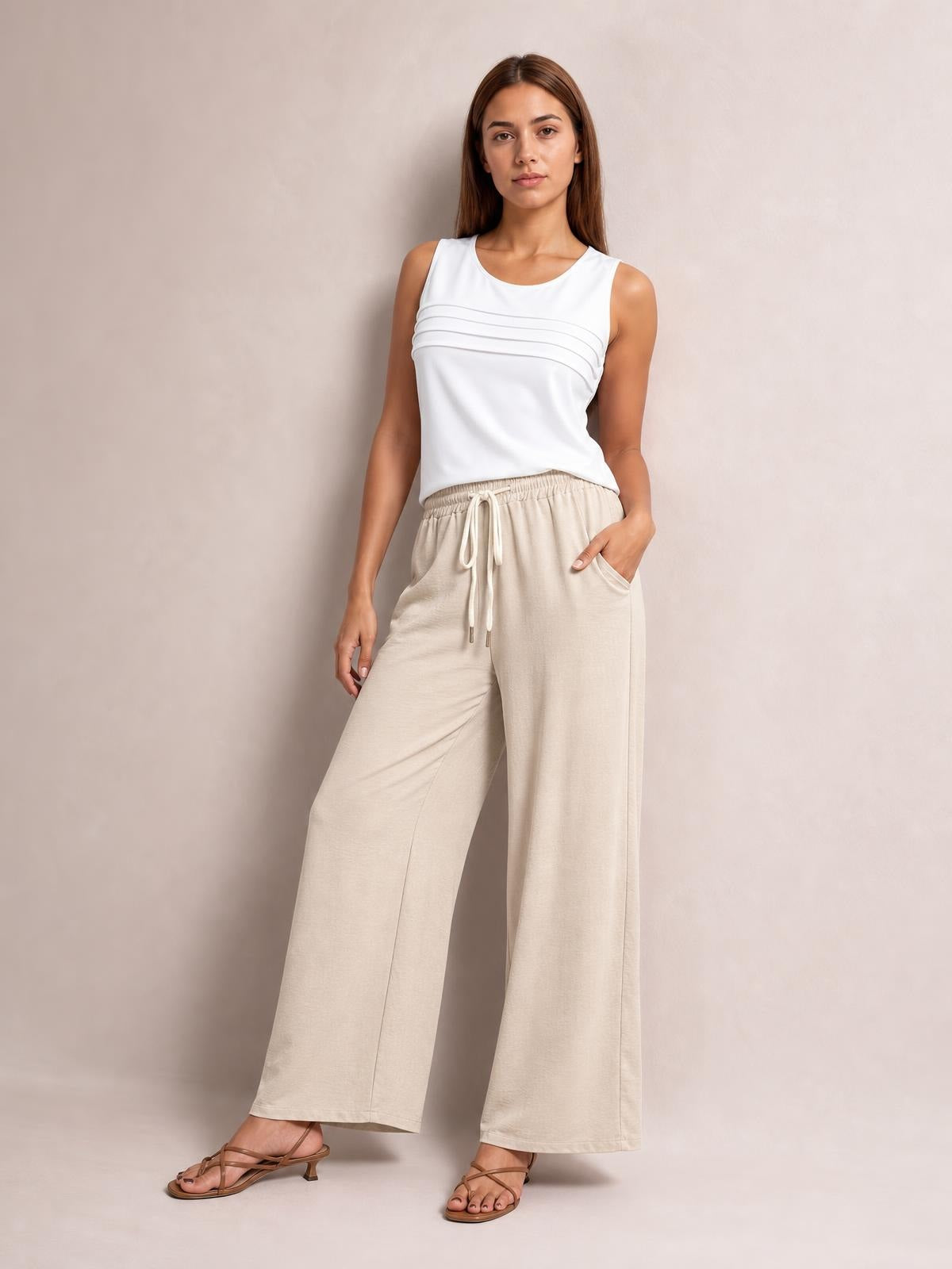 Model wears beige drawstring wide leg pants with an elastic waist, visible front pockets, and a relaxed full-length silhouette while standing in strappy sandals.