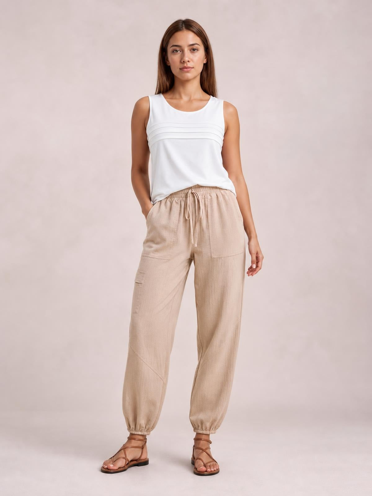 Model stands in a relaxed pose wearing beige elastic-waist joggers with a drawstring, multiple pockets, a loose tapered silhouette, and gathered ankle cuffs.