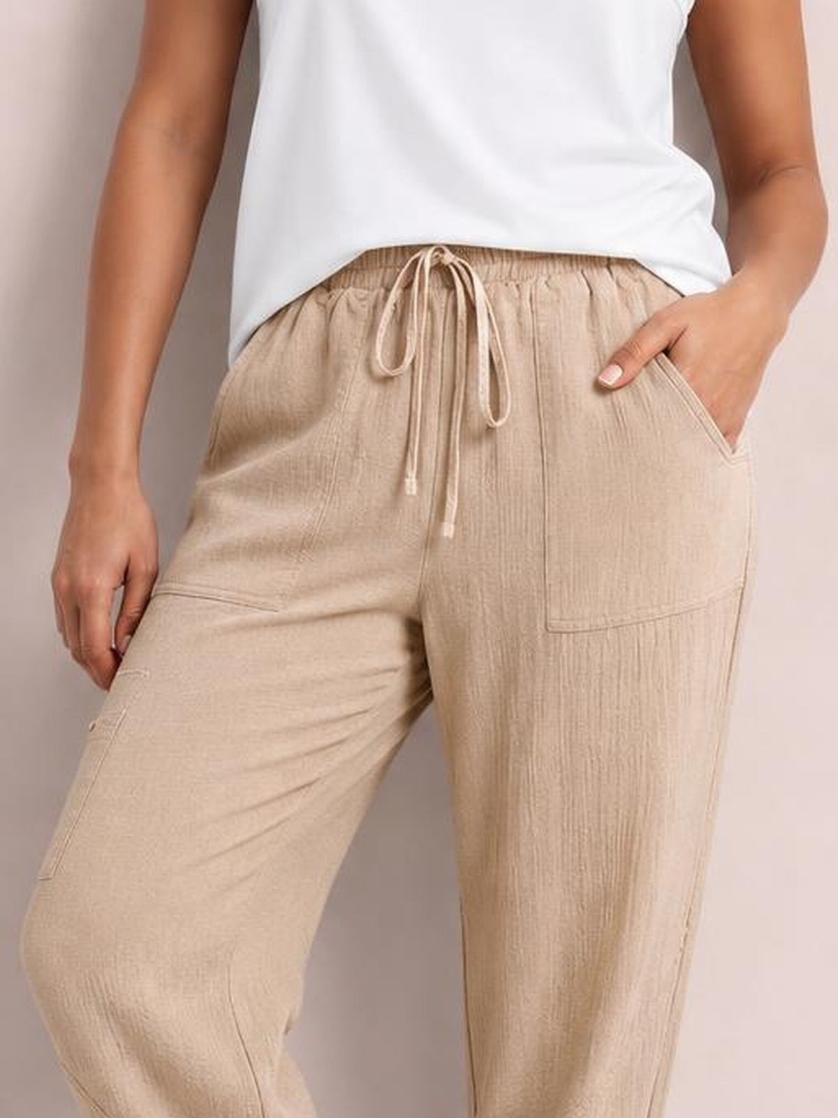 A model wears beige elastic waist joggers with a drawstring, relaxed straight-leg silhouette, and practical side pockets, with one hand tucked inside.