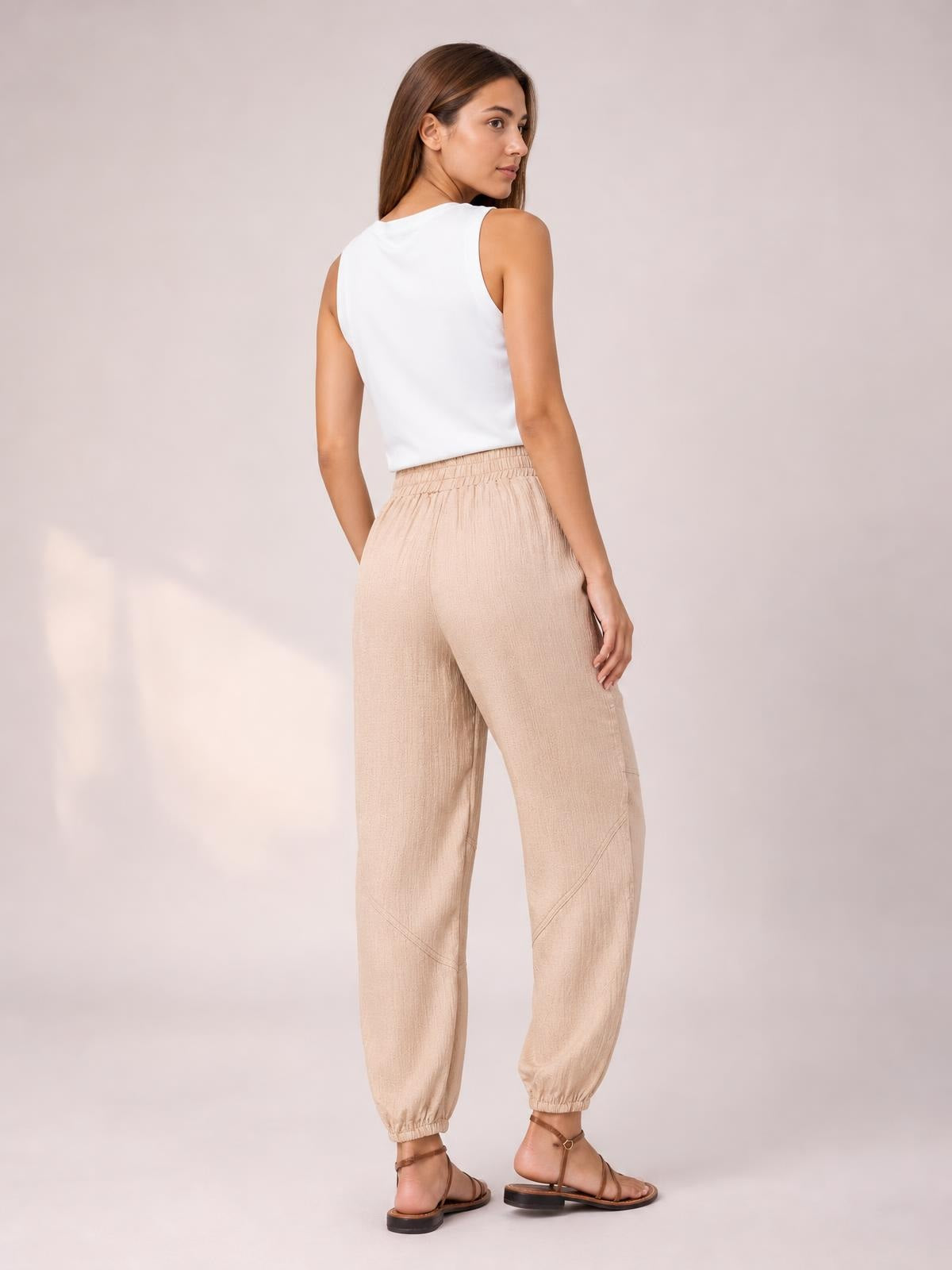 A model poses turned away in beige elastic-waist joggers with a relaxed tapered silhouette, side pockets, and gathered ankle cuffs.