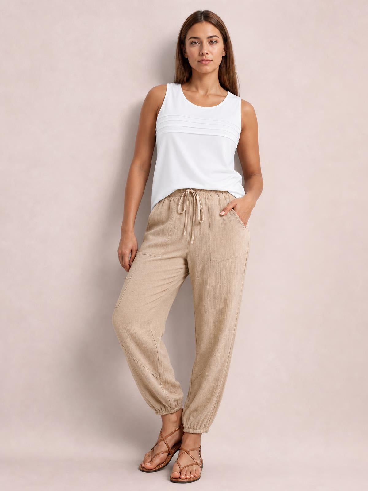 Model wearing beige elastic waist joggers with a drawstring, roomy pockets, relaxed tapered legs, and elastic ankle cuffs, posed with one hand in a pocket.