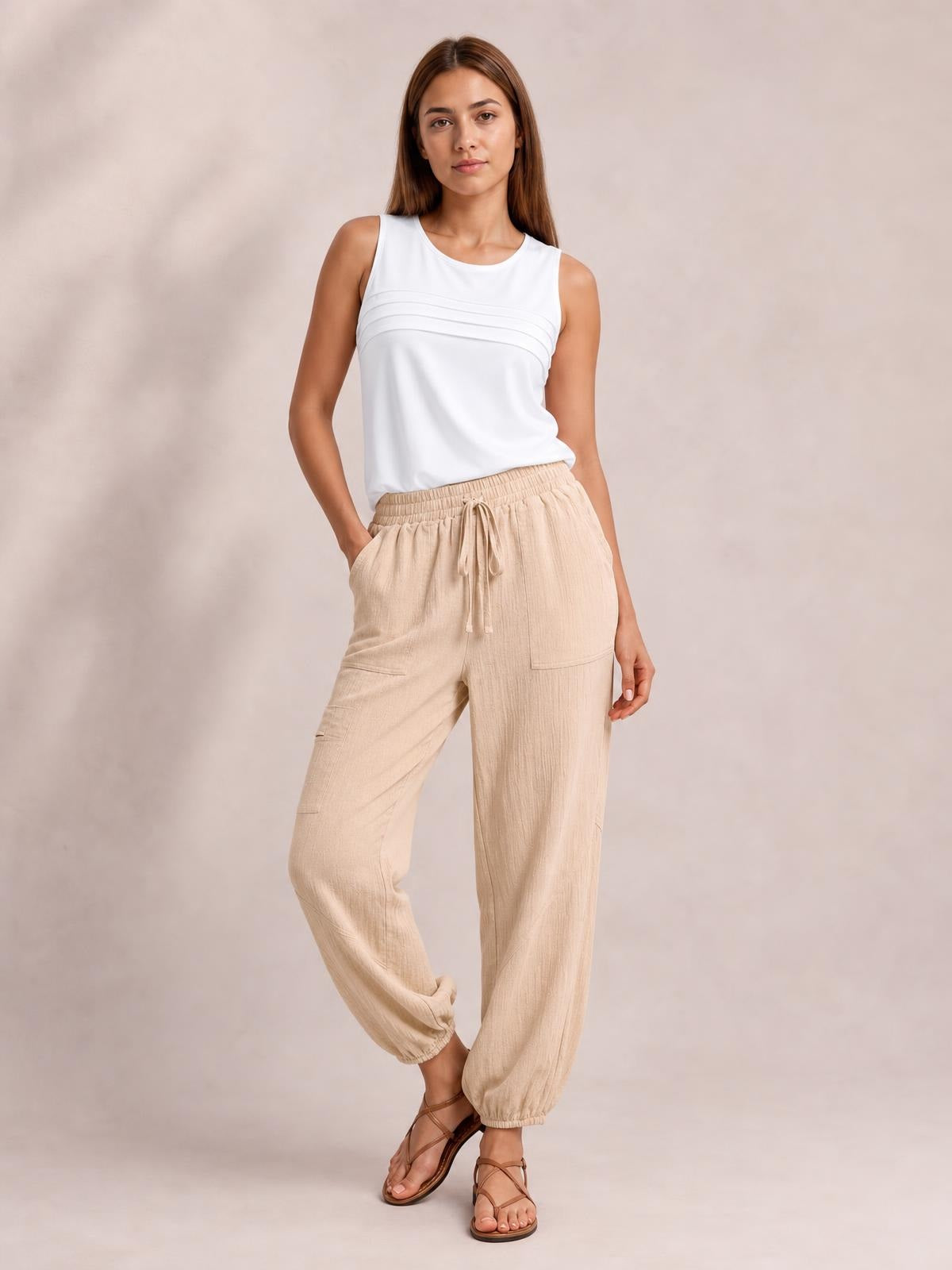A model poses in beige elastic-waist joggers with a drawstring, roomy pockets, relaxed tapered legs, and gathered ankle cuffs, one hand in a pocket.
