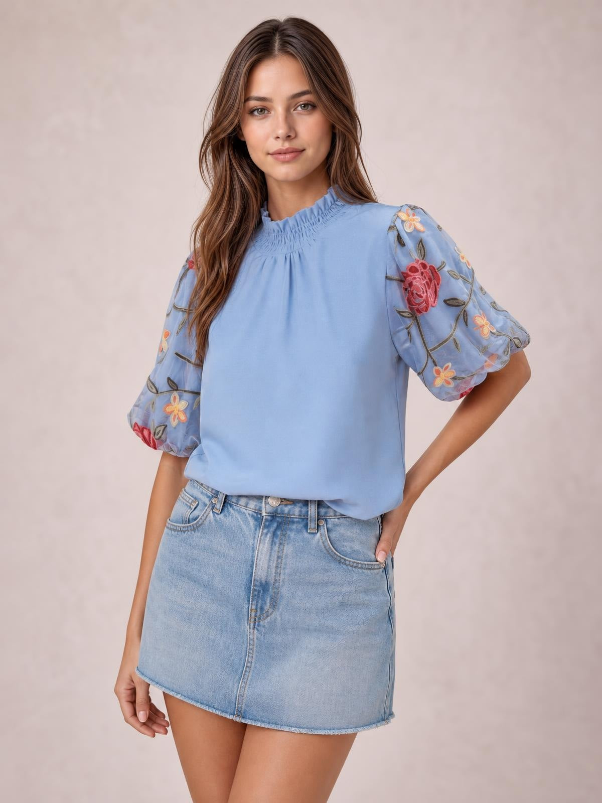 A model poses in a light blue embroidered mock neck blouse with voluminous puff sleeves, gathered neckline, and a relaxed silhouette.