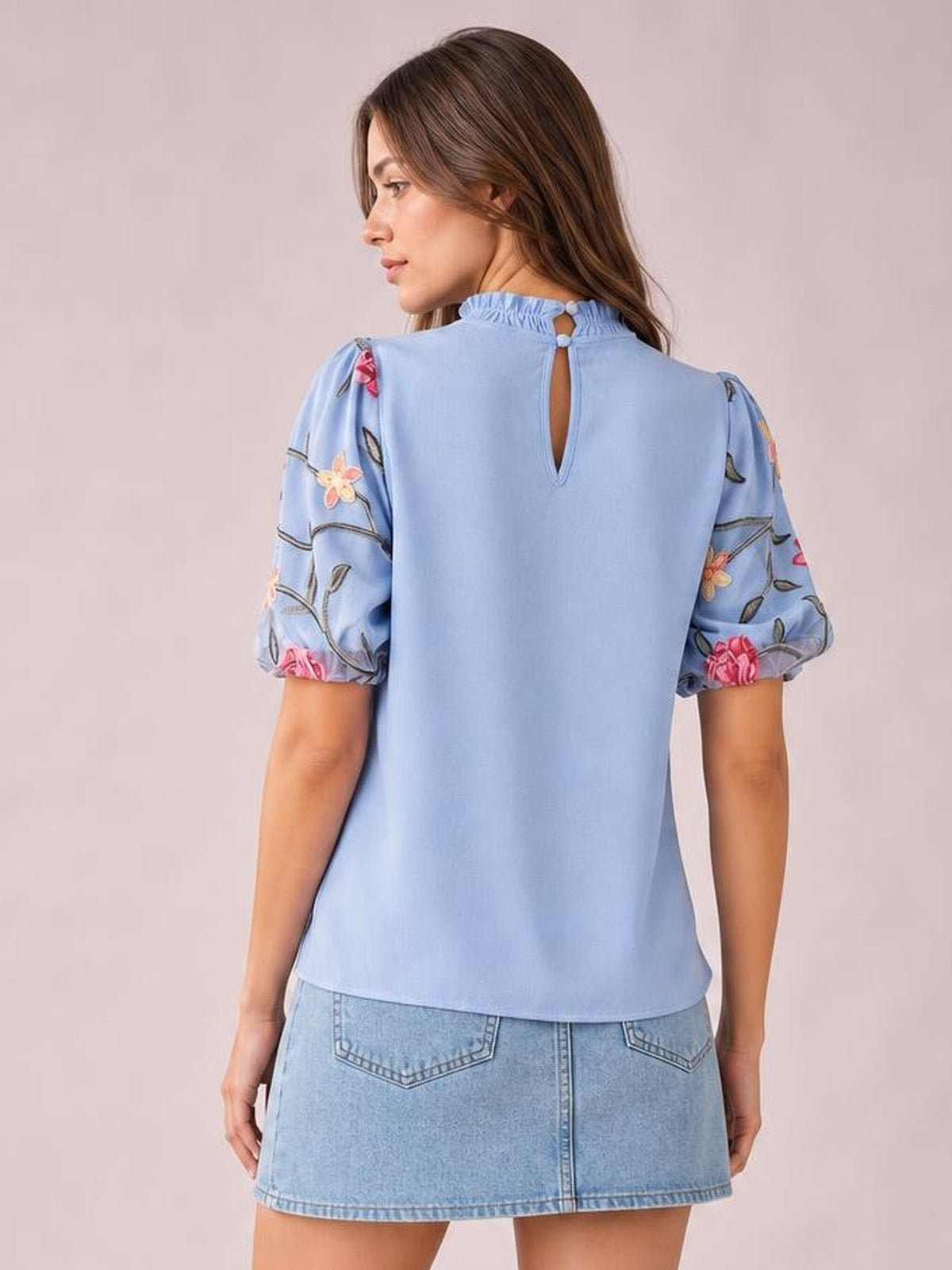 Model shown from behind in a light blue embroidered mock neck blouse with puff sleeves, floral detailing, buttoned keyhole back, and a relaxed straight silhouette.