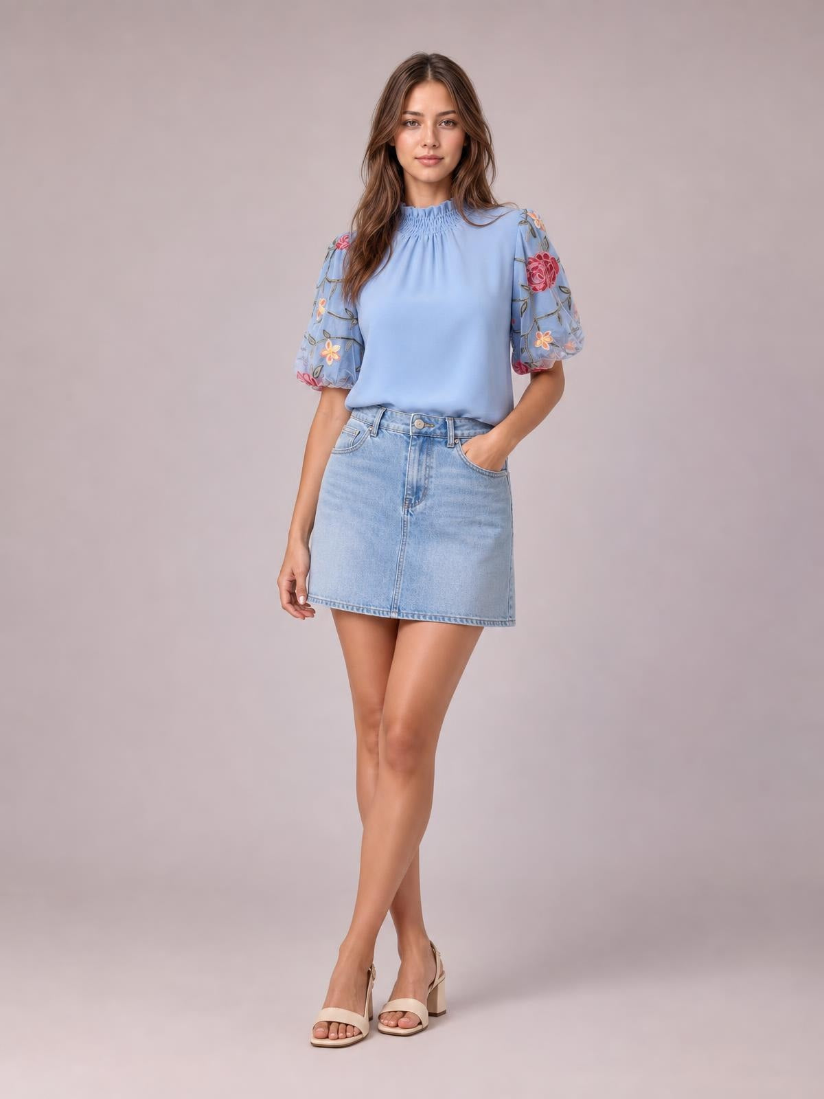 A model stands with one hand in her pocket wearing a light blue embroidered mock neck blouse with puff sleeves and a relaxed silhouette.