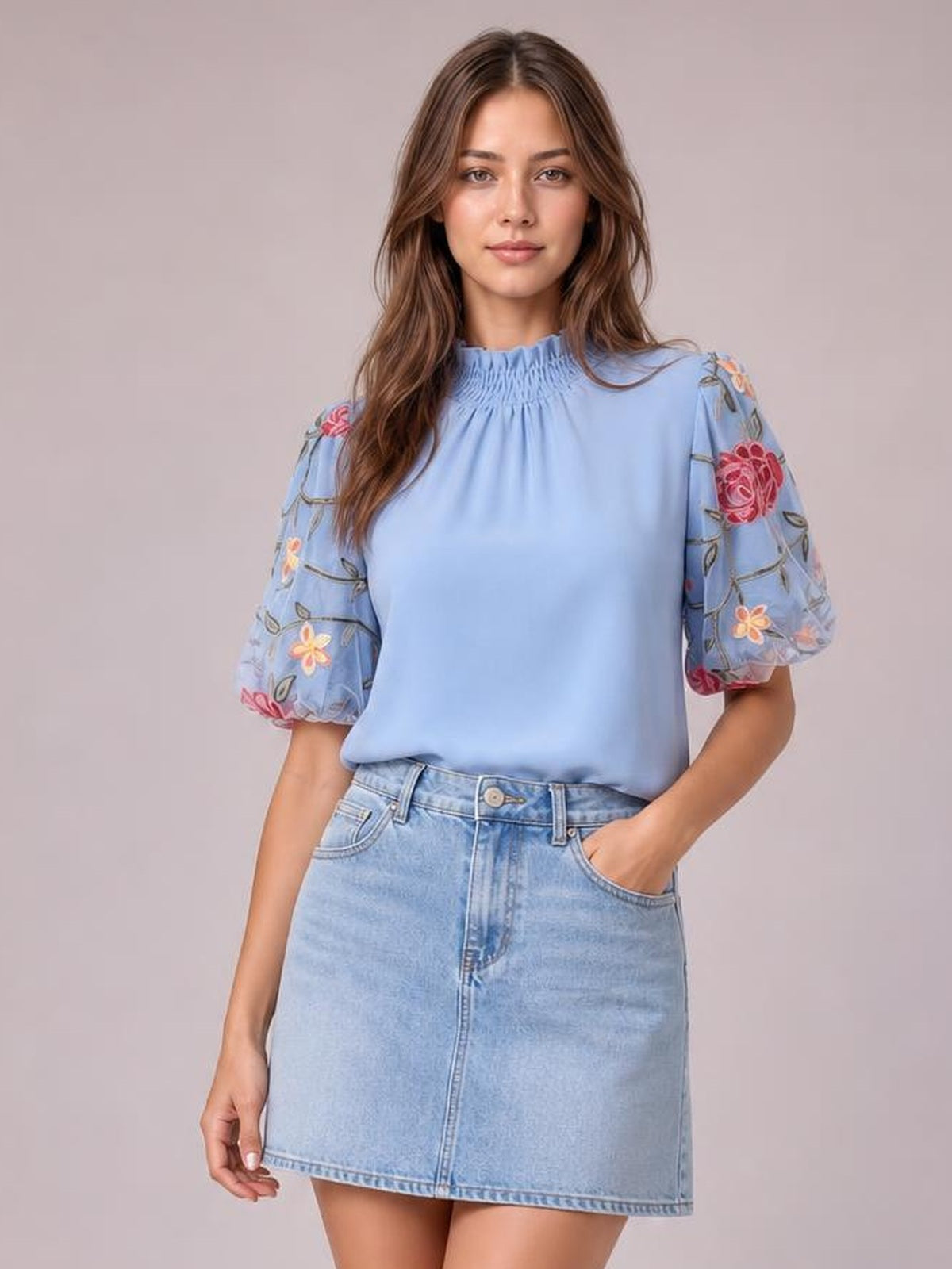 Model poses with one hand in her pocket wearing a light blue embroidered mock neck blouse with sheer floral puff sleeves and a relaxed silhouette.