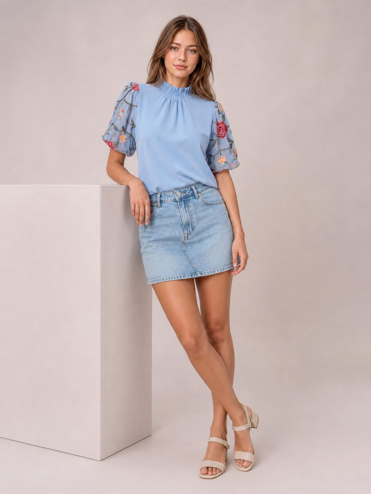 A model wears a light blue embroidered mock neck blouse with gathered neckline, puff sleeves, and a relaxed silhouette while leaning with one hand resting on a pedestal.