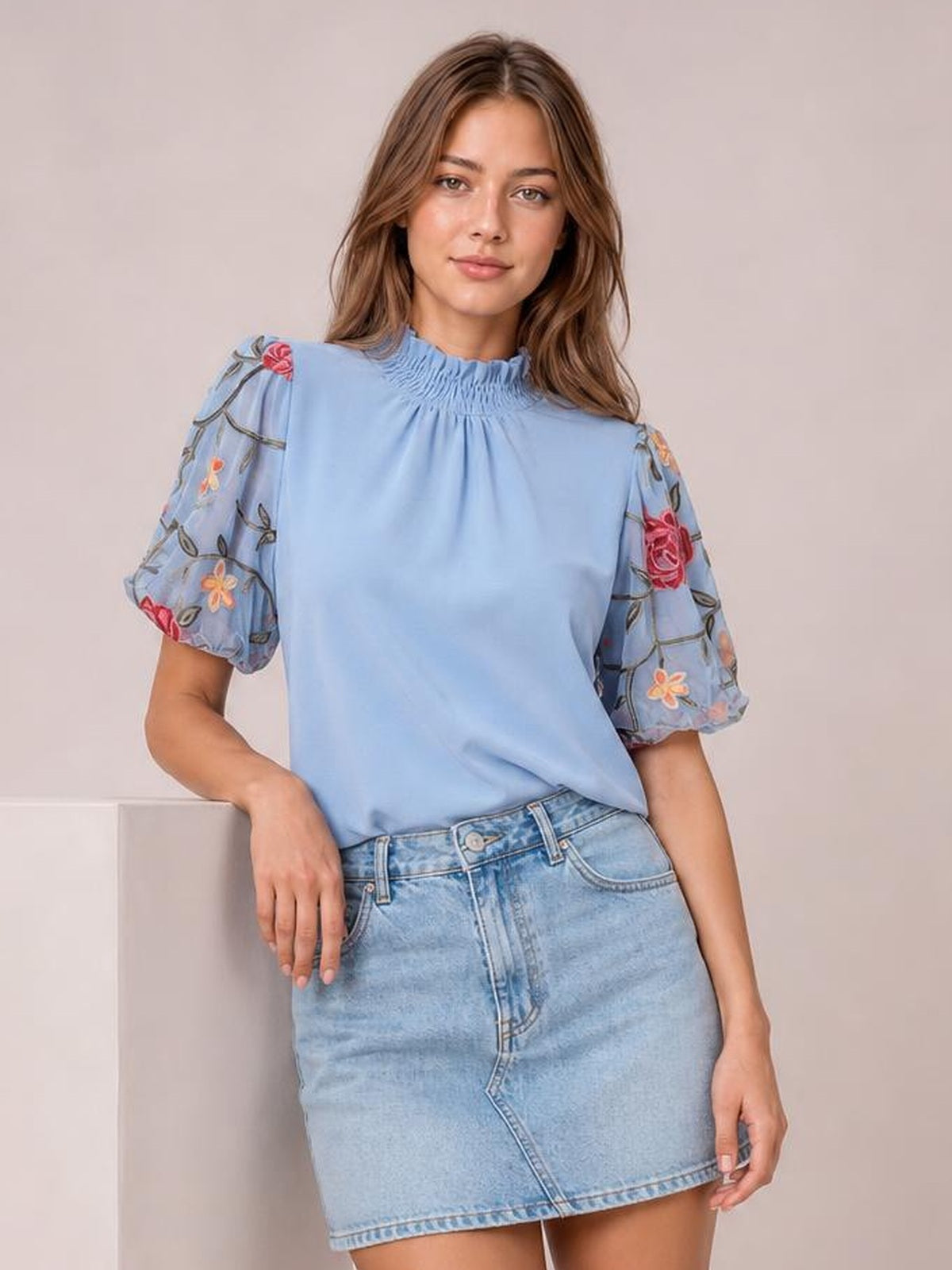A model poses in a light blue embroidered mock neck blouse with a softly gathered neckline, puff sleeves, and a relaxed silhouette.