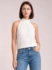 A model wears a white eyelet Grecian neck tank with a high neckline, sleeveless cut, and relaxed straight silhouette, posing with one hand in her jeans pocket.