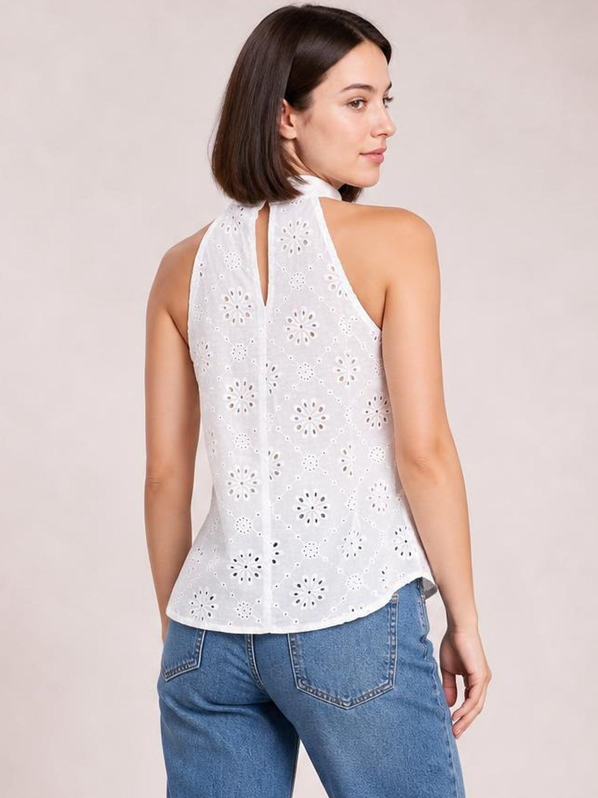 A model looks over her shoulder in a white eyelet Grecian neck tank with a halter collar, back keyhole, sleeveless cut, and gently flared silhouette.