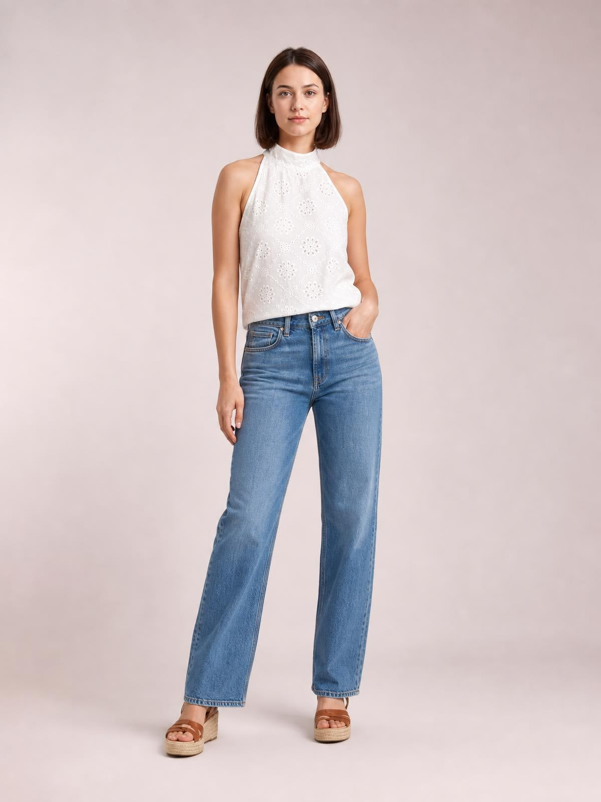 Model wearing a white eyelet Grecian neck tank with a high neckline, sleeveless cut, floral perforations, and a relaxed silhouette, posing with one hand in her jeans pocket.