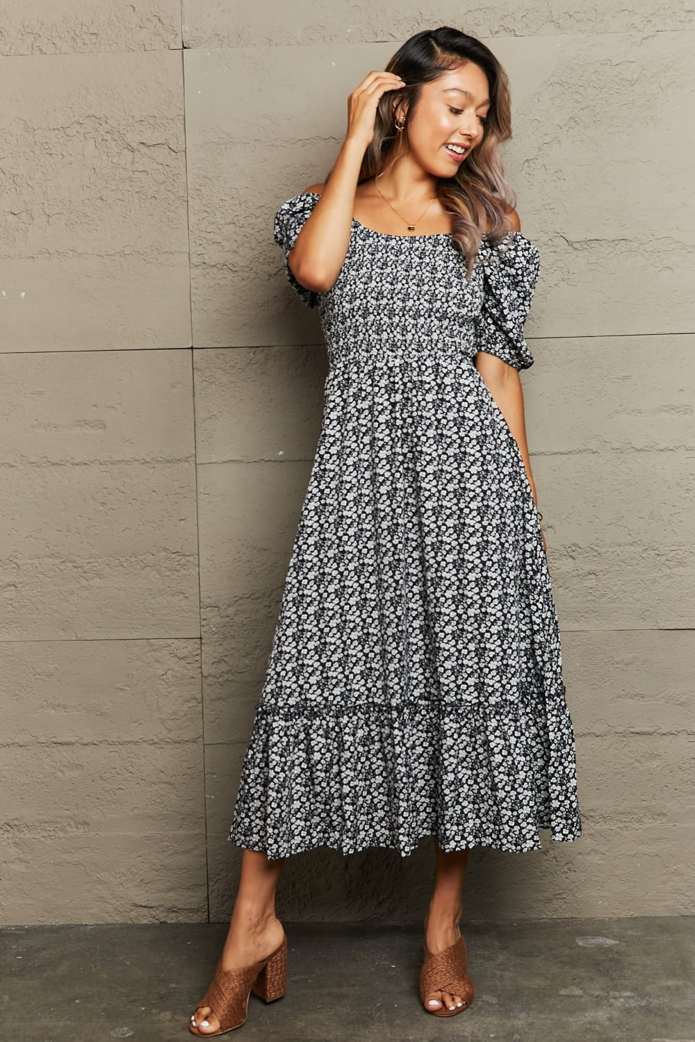 A model poses while touching her hair in a black floral midi dress with an off-shoulder square neckline, short puff sleeves, gathered A-line shape, and ruffled hem.
