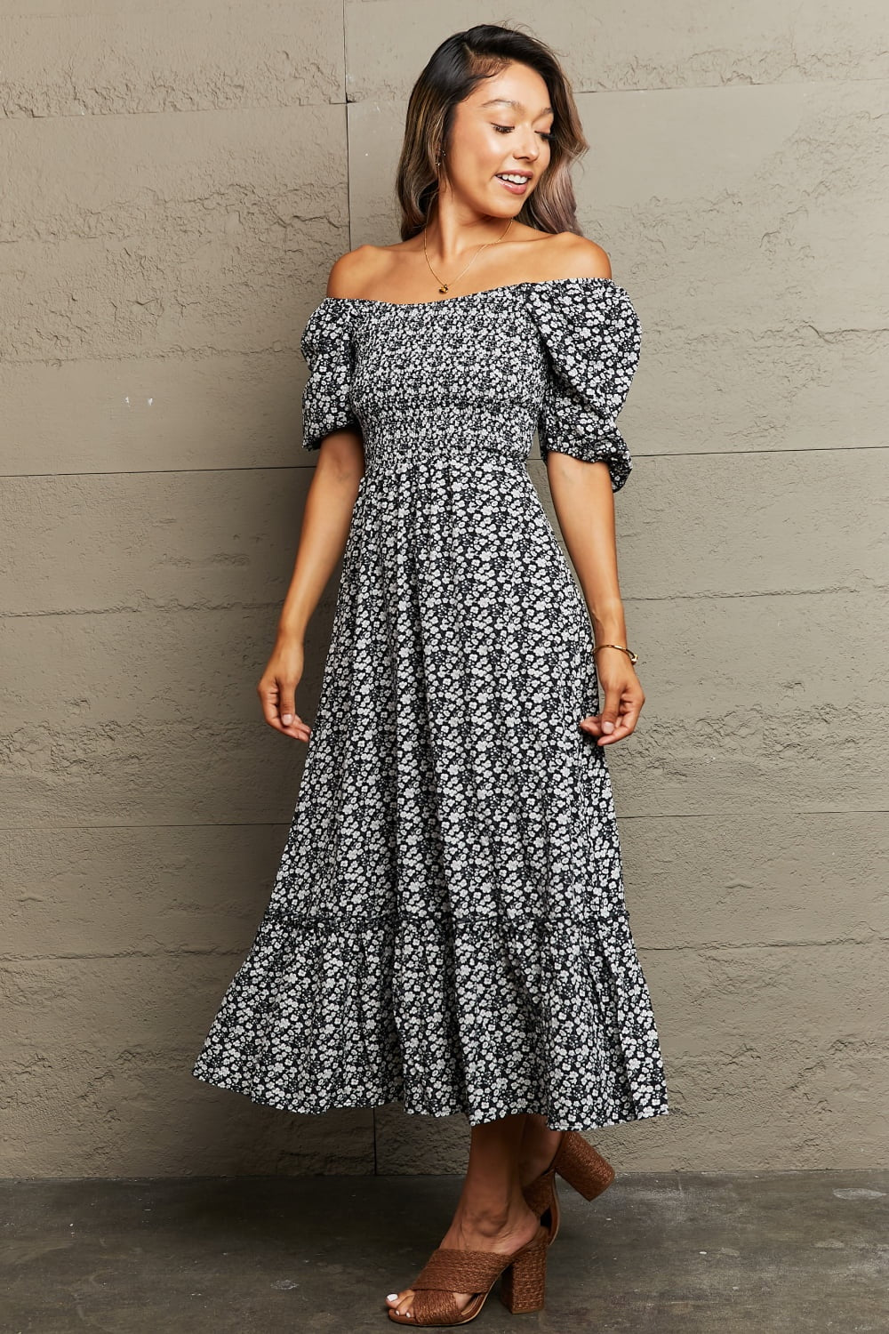 Model wears a black floral off-shoulder midi dress with puff sleeves, a gathered A-line silhouette, ruffle hem, and crossed ankles.