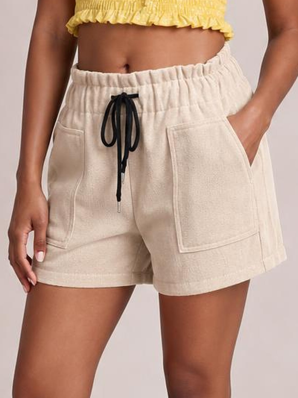 Model wears cream drawstring shorts with an elastic waistband, black tie, large front pockets, and a relaxed straight-leg silhouette.
