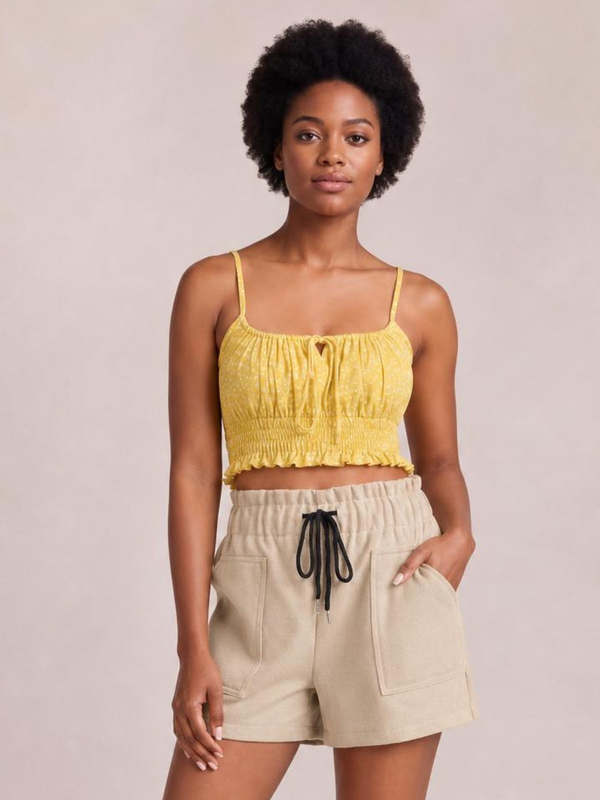 A woman modeling a yellow textured smocked crop top with thin straps and beige drawstring shorts against a solid light beige background.