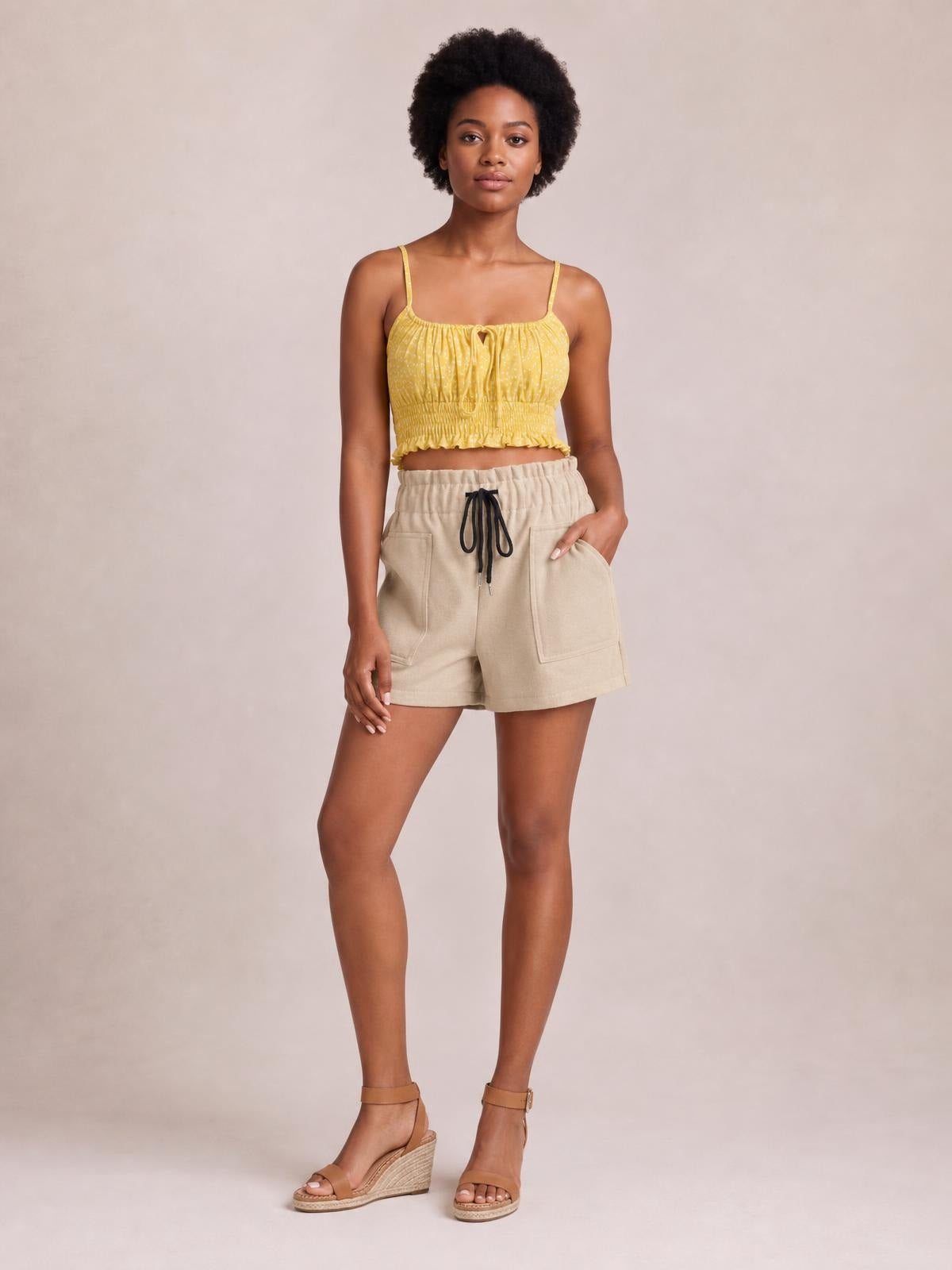 A model stands with one hand in the pocket, wearing beige drawstring shorts with front patch pockets and a relaxed mid-thigh silhouette.