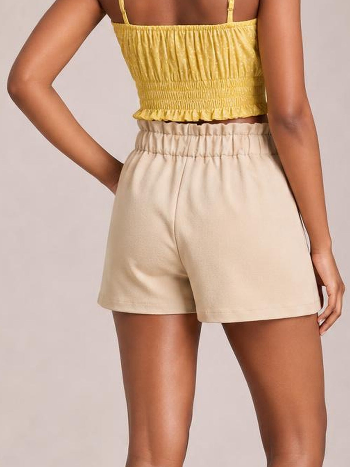 A model wears beige shorts with a high elasticated waistband, relaxed silhouette, and side pocket, posing with one hand tucked inside.