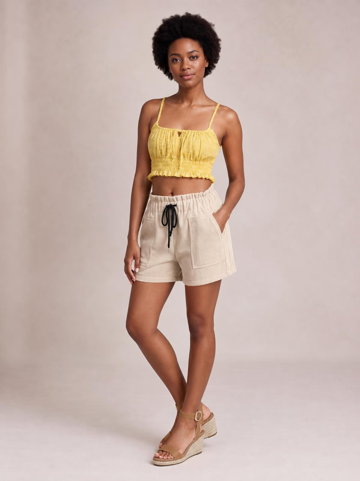 A model poses in relaxed beige drawstring shorts with an elastic waist, loose silhouette, and large front patch pockets, with one hand tucked inside.
