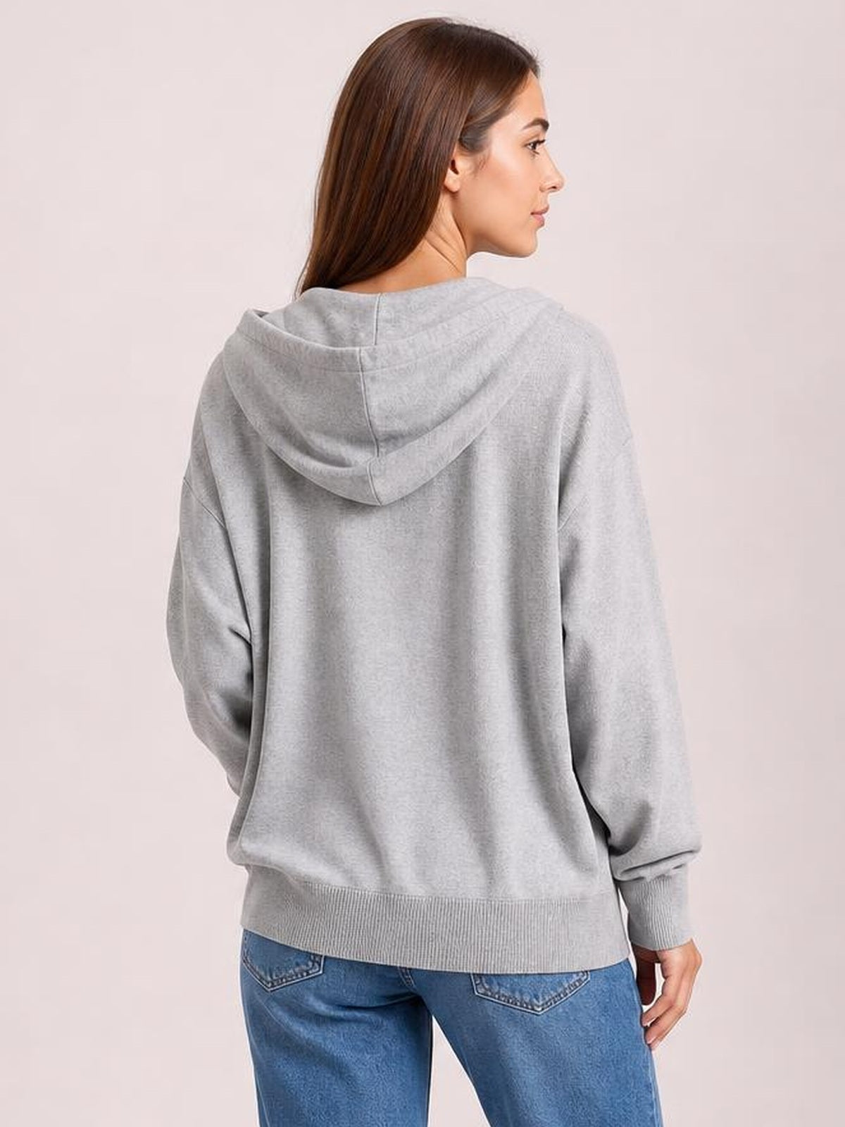 A model turns to show the back of a light gray long-sleeve hoodie with a roomy hood, relaxed silhouette, ribbed cuffs, and hem.