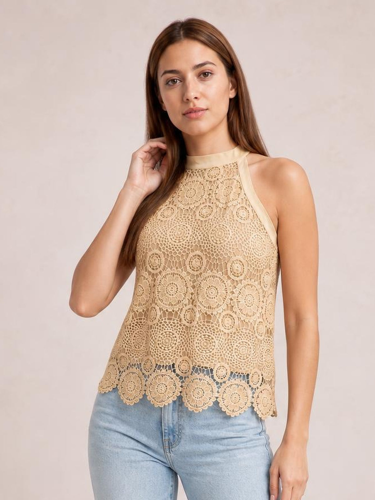 Model wearing a beige lace mock neck tank with a sleeveless silhouette, intricate circular pattern, and scalloped hem, posing with one hand near her hair.