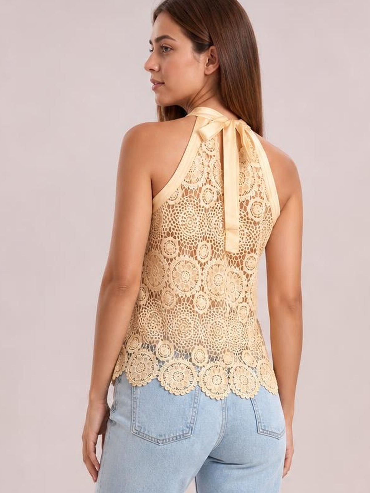 Back view of a model wearing a cream lace mock neck tank with a tied neck bow, relaxed silhouette, and scalloped hem over light-wash jeans.