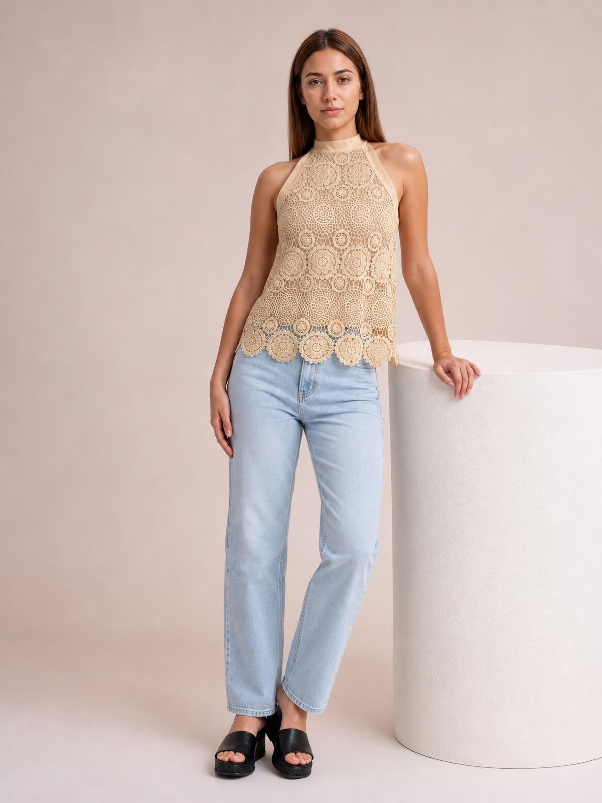 Model wears a beige lace mock neck tank with sleeveless halter styling, a relaxed straight silhouette, and scalloped hem while leaning on a pedestal.