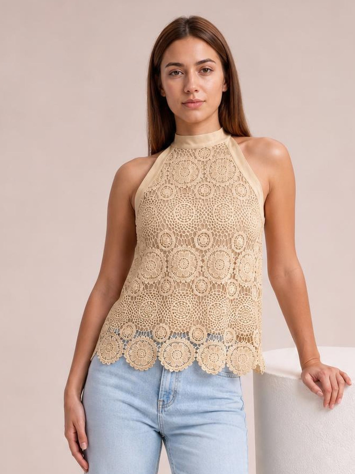A model stands wearing a cream lace mock-neck tank with halter-style straps, a relaxed straight silhouette, and a scalloped floral hem.