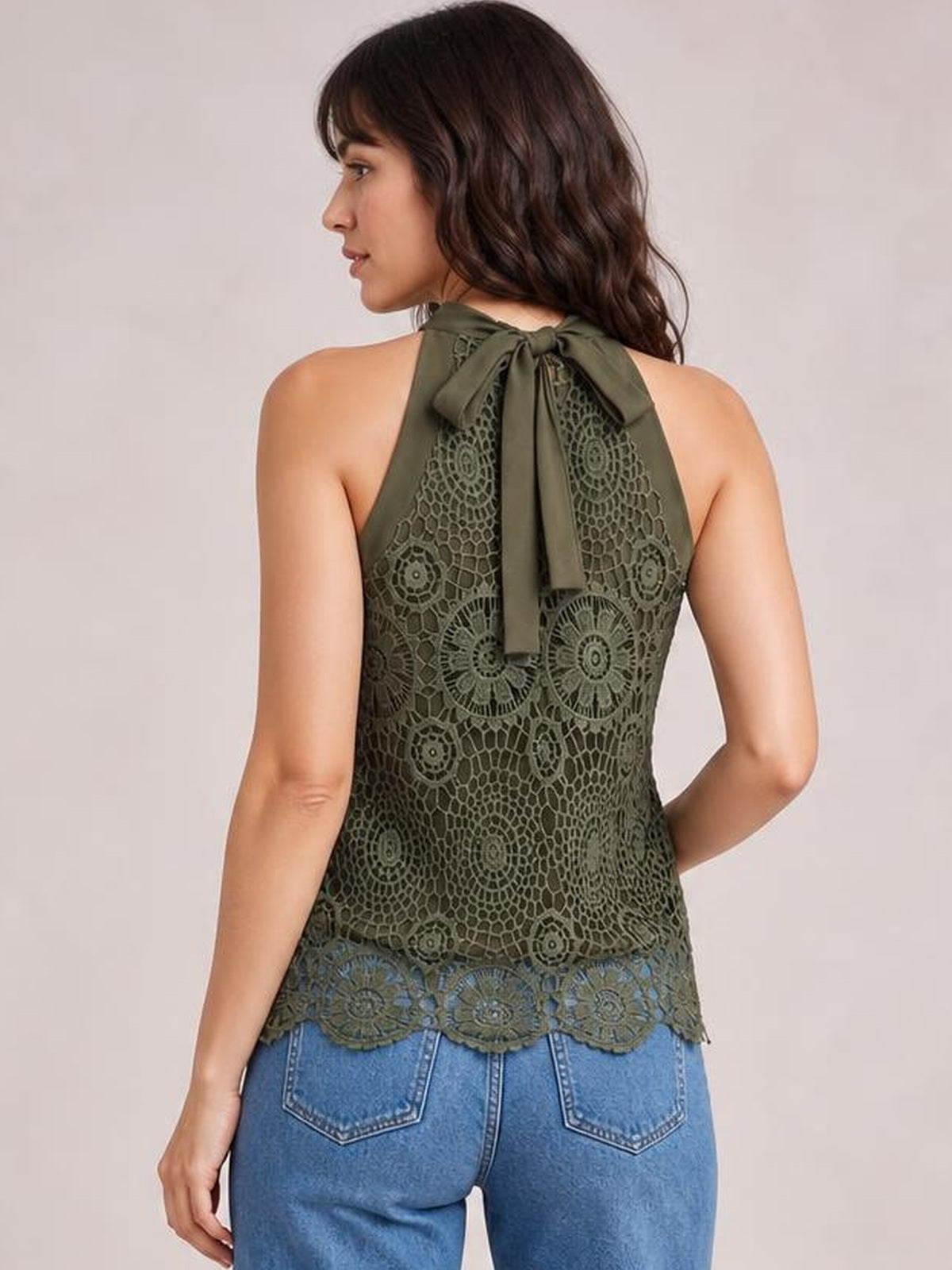 Model poses turned away in an olive lace mock neck tank with a tied halter neckline, intricate floral lace pattern, and scalloped hem.