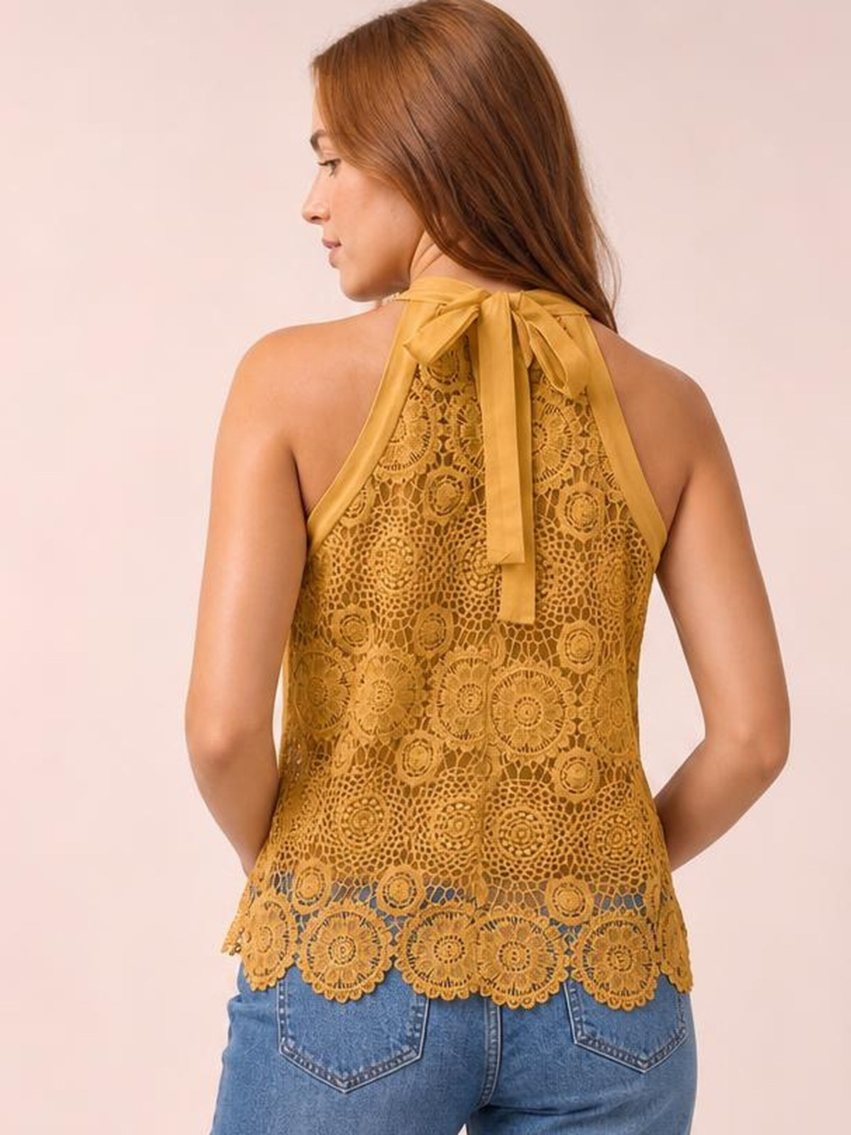 A model looks over her shoulder in a mustard lace tank with a tied mock neck, halter-style straps, relaxed silhouette, and scalloped hem.