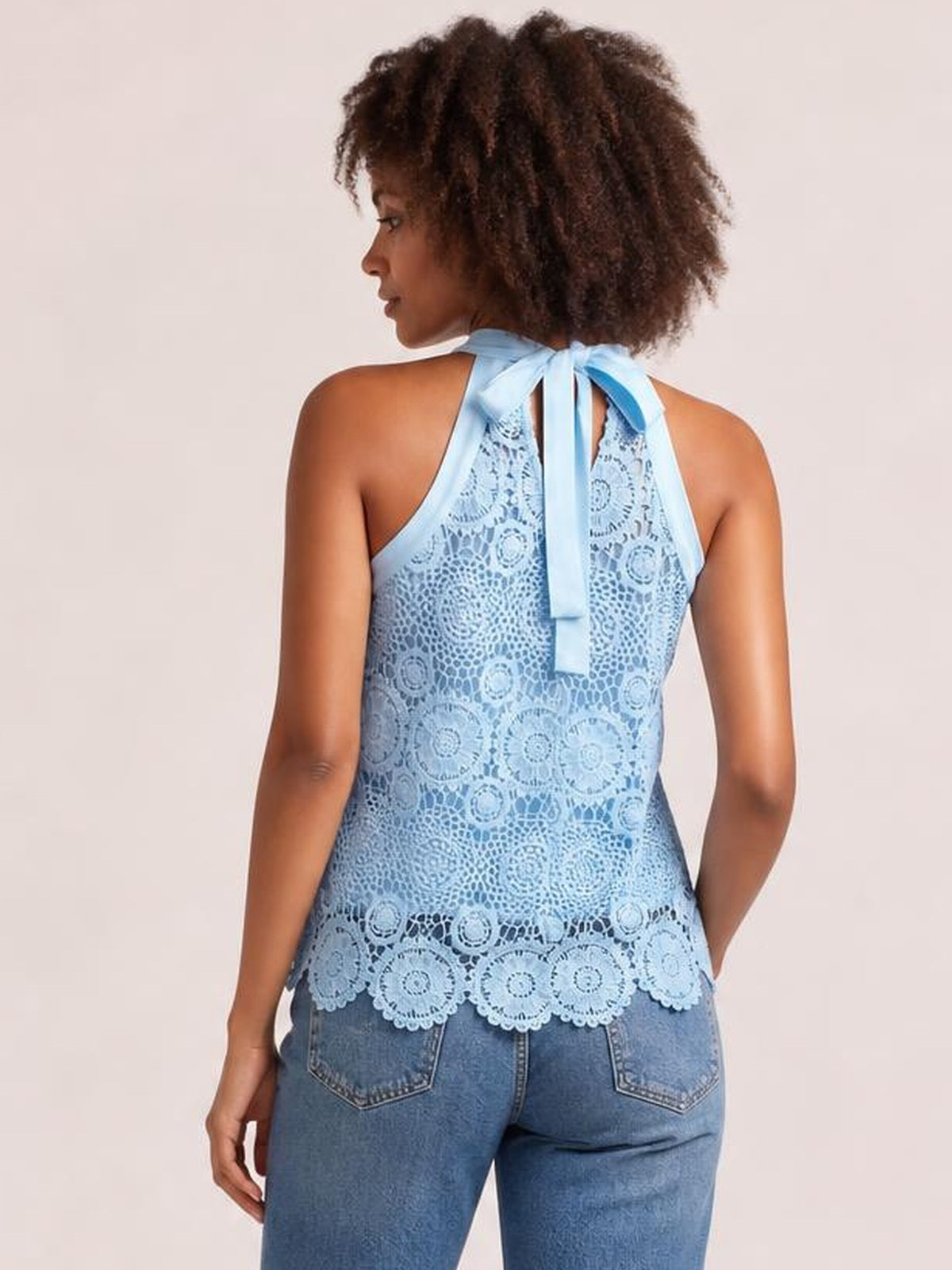 A model wears a light blue lace tank with a tied mock neck, halter-style straps, and scalloped hem, shown from the back in a relaxed pose.