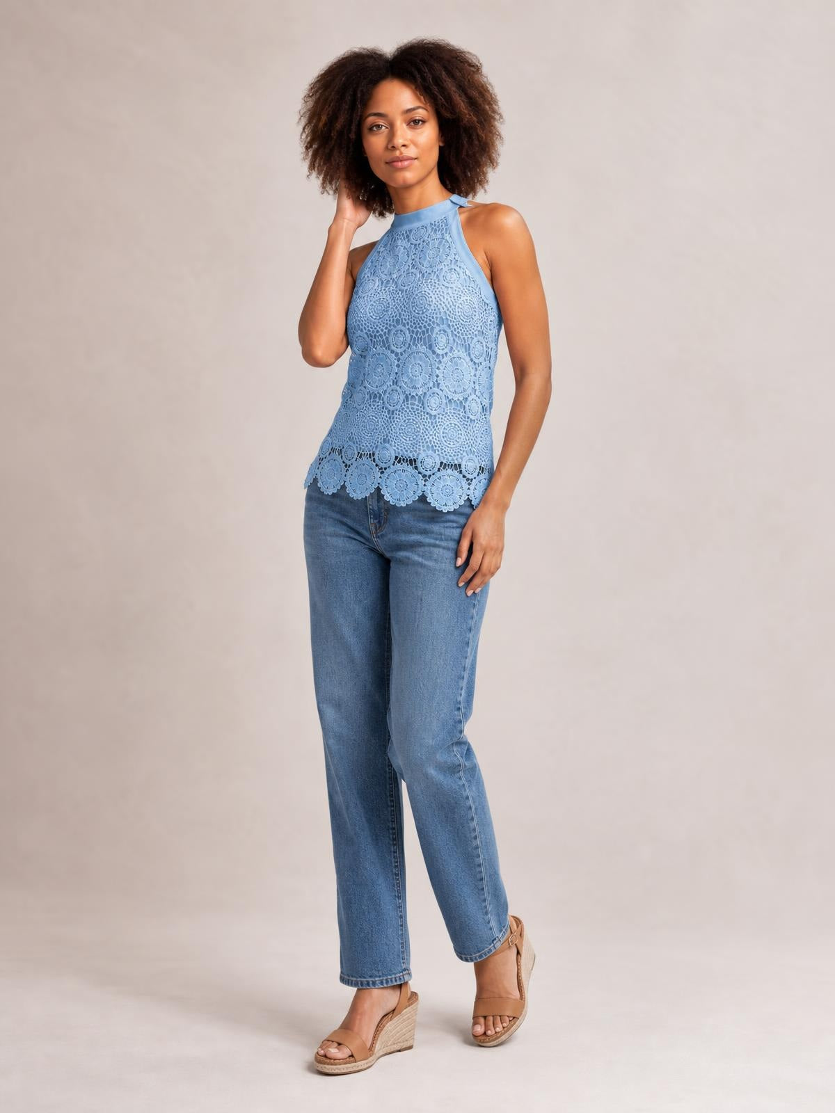 A model poses in a light blue lace mock neck tank with a fitted sleeveless silhouette, halter-style straps, and scalloped hem while touching her hair.