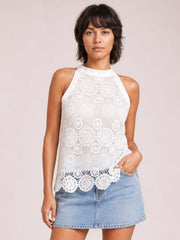 Model wears a white lace mock neck tank with sleeveless cut, relaxed silhouette, intricate floral construction, and scalloped hem, posing with one hand in her pocket.