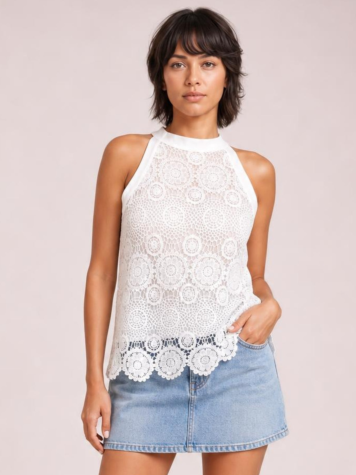 Model wears a white lace mock neck tank with sleeveless cut, relaxed silhouette, intricate floral construction, and scalloped hem, posing with one hand in her pocket.