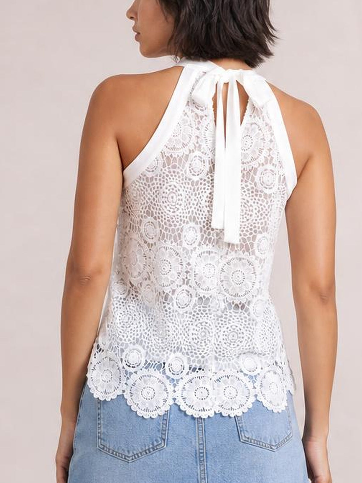 Model viewed from the back wears a white lace tank with an opaque lining, mock neck, tied halter-style neckline, relaxed silhouette, and scalloped hem.