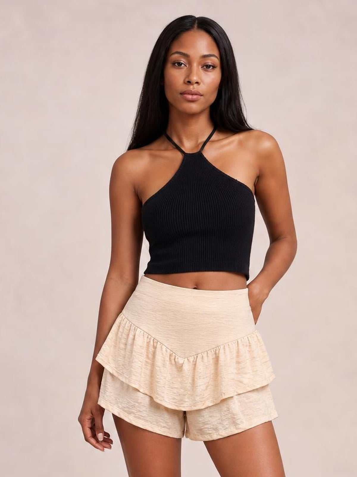 A woman modeling a black ribbed halter crop top and cream ruffled shorts against a solid beige studio background.