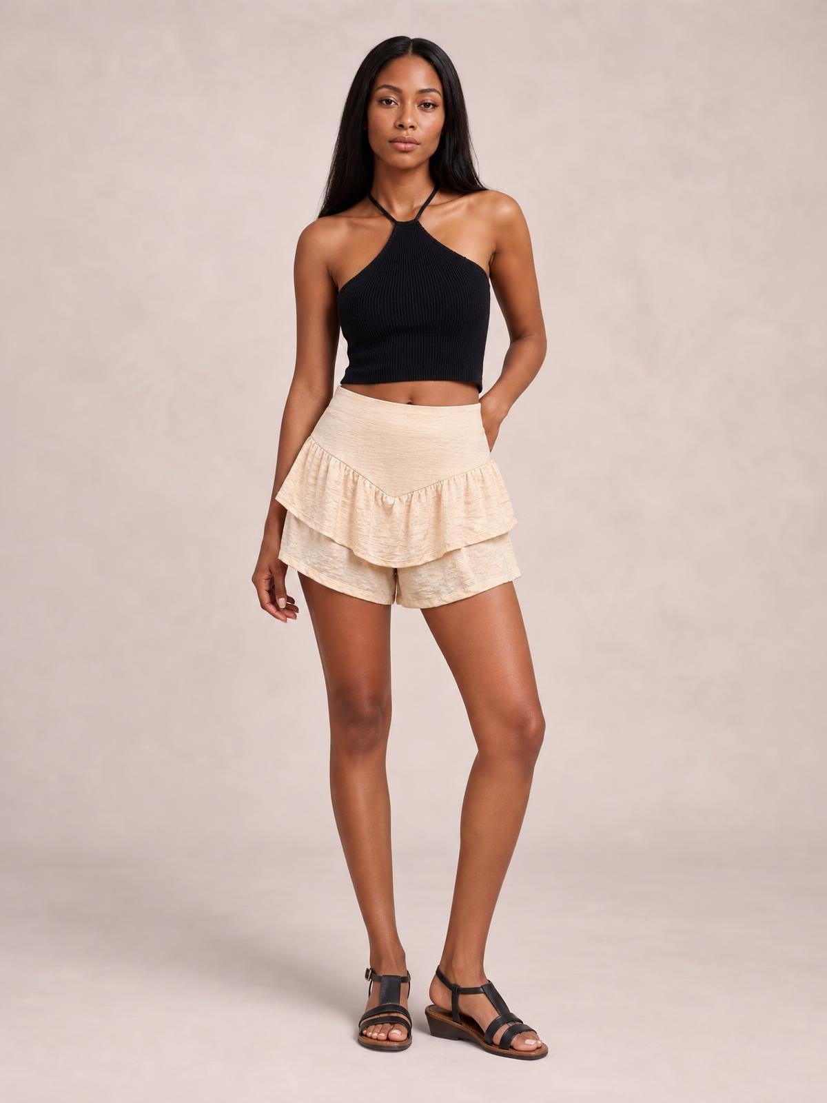 A model poses with one hand in a pocket, wearing cream high-waist layered shorts with a wide waistband, tiered ruffles, and a relaxed silhouette.