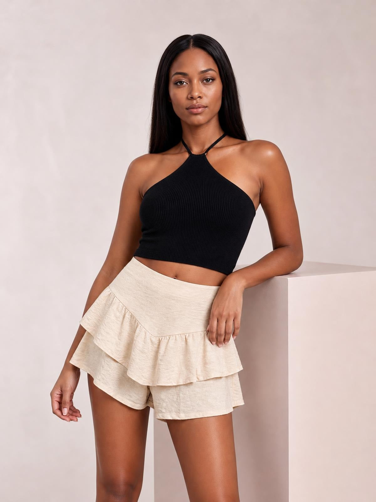A model leans against a ledge wearing cream high-waisted layered shorts with tiered ruffle panels and a relaxed silhouette.