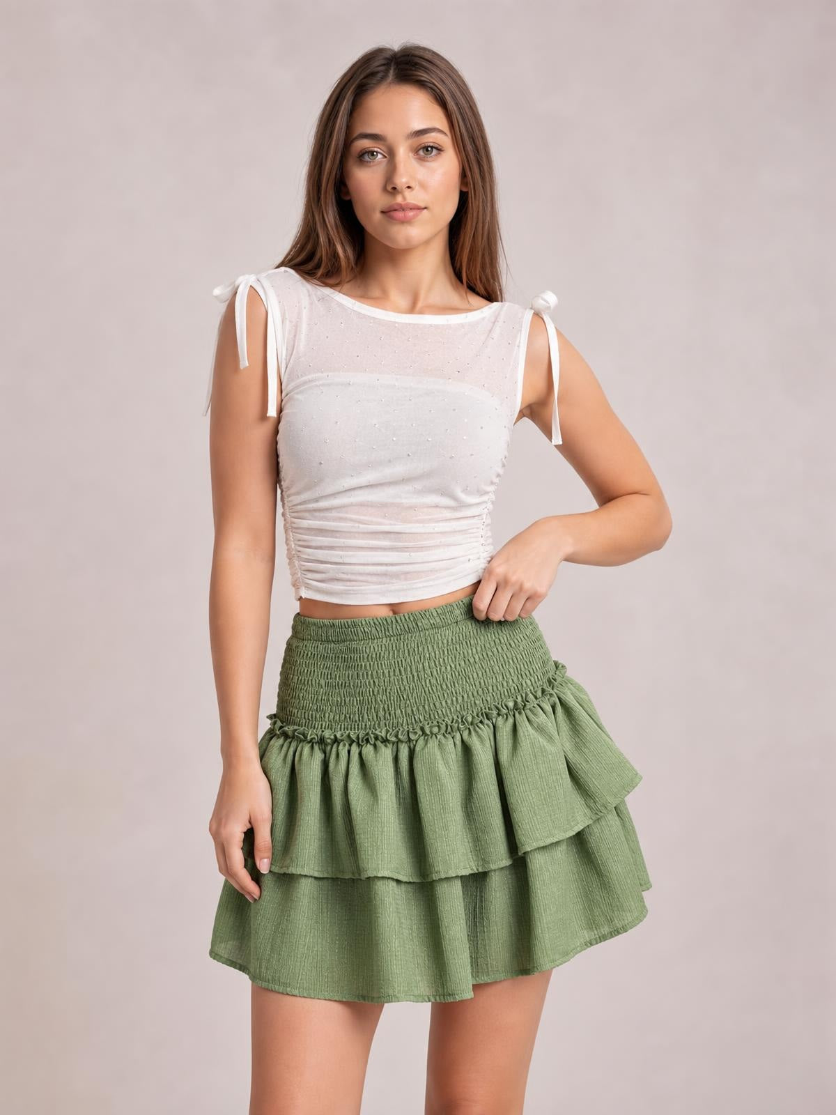 Model poses in a sage green smocked mini skirt with a tiered layered silhouette, gathered waistband, and ruffled frill trim.
