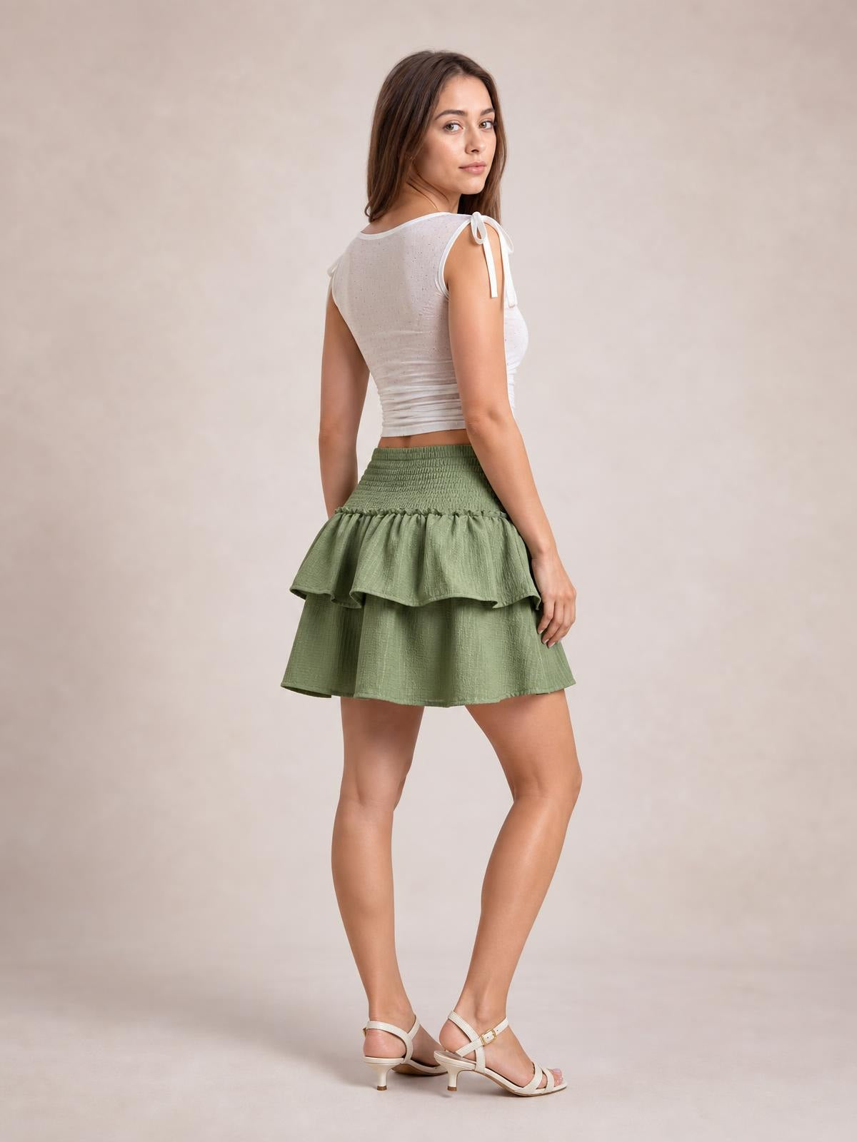 Model poses in a sage green layered smocked mini skirt with a flounced frill tier and gently flared silhouette.