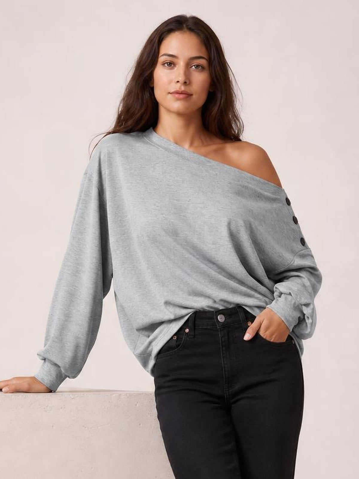 Model poses in a gray off-shoulder long-sleeve blouse with a relaxed silhouette, button detailing along the side, and one hand in her jeans pocket.