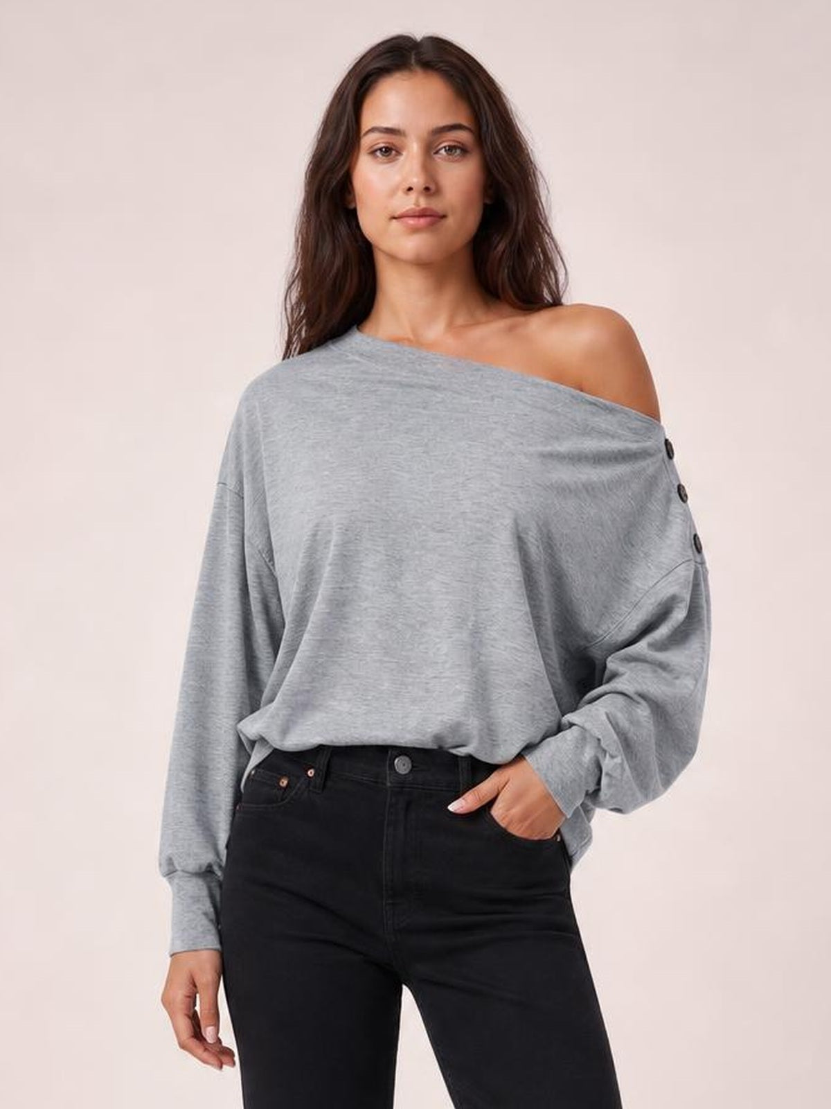 A model wears a solid gray off-shoulder long-sleeve blouse with a relaxed silhouette, button accents along the sleeve, and one hand resting in her jeans pocket.