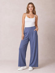 Model wearing solid blue pleated-detail wide-leg pants with a high waistband and side pockets, posed with one hand tucked into a pocket.