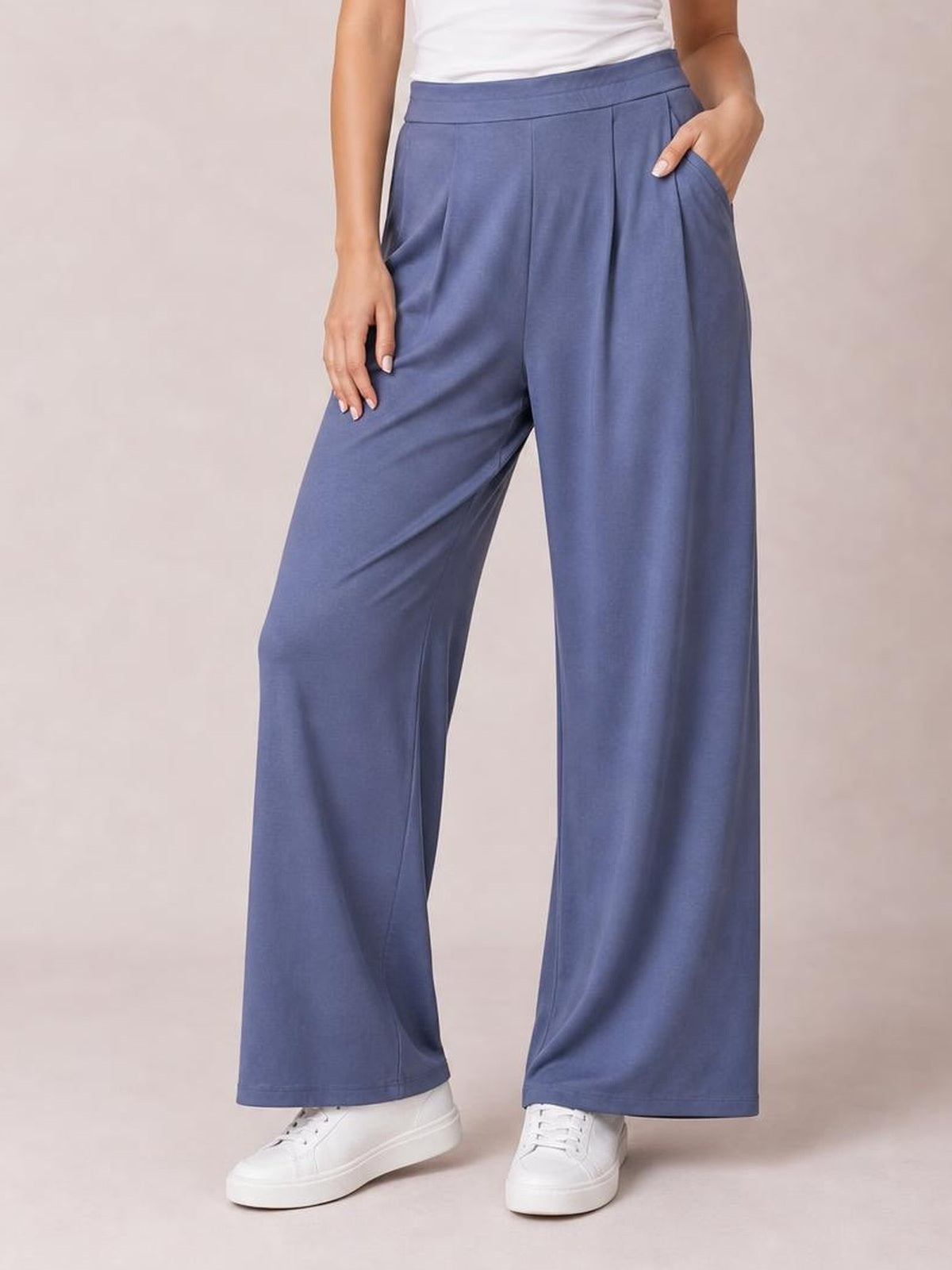 Model wearing blue pleated-detail wide-leg pants with a high waistband and side pockets, standing in white sneakers with one hand in a pocket.