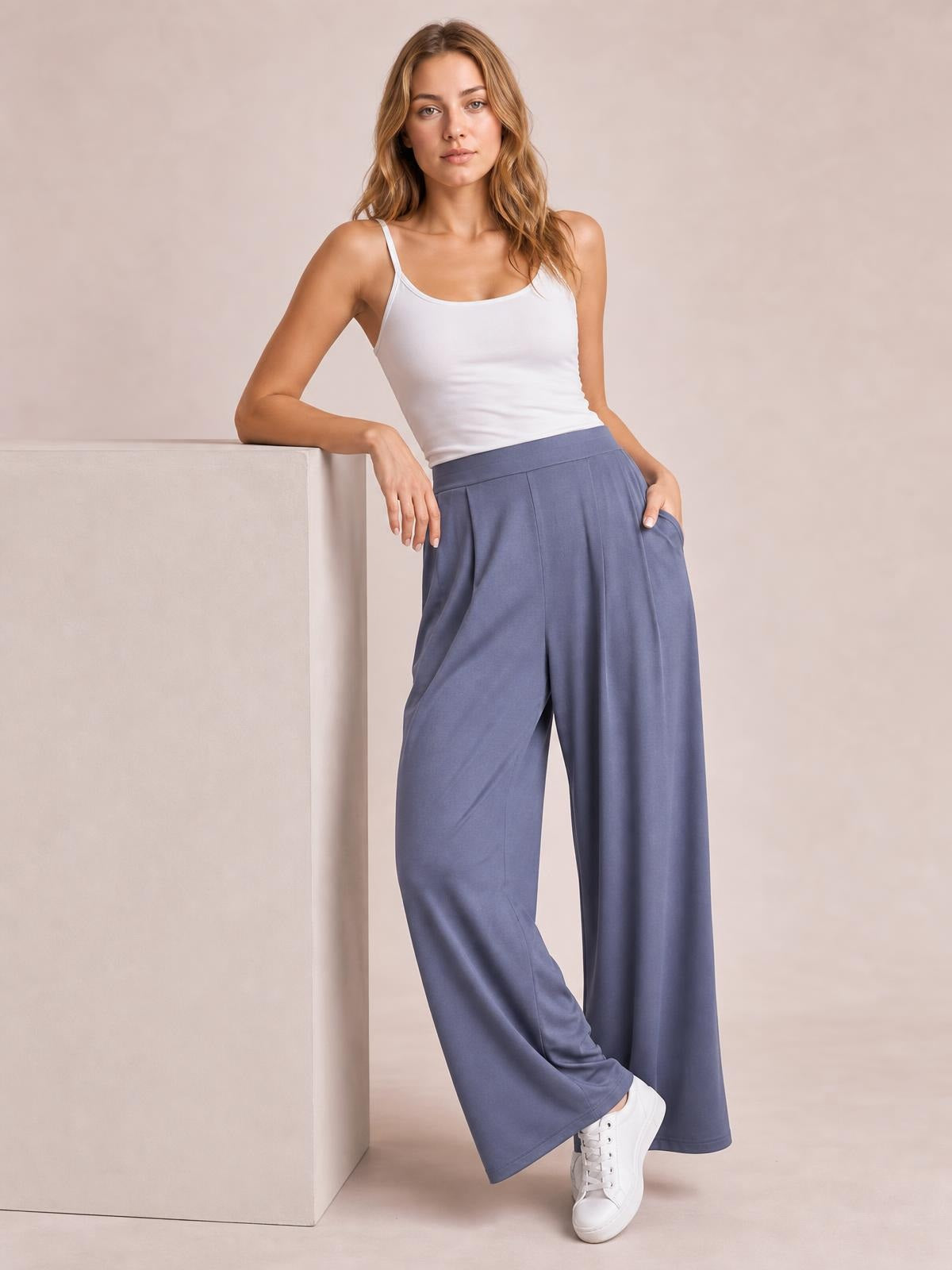 Model leans against a block wearing solid blue pleated-detail wide-leg pants with a high waistband, long flowing silhouette, and side pockets.