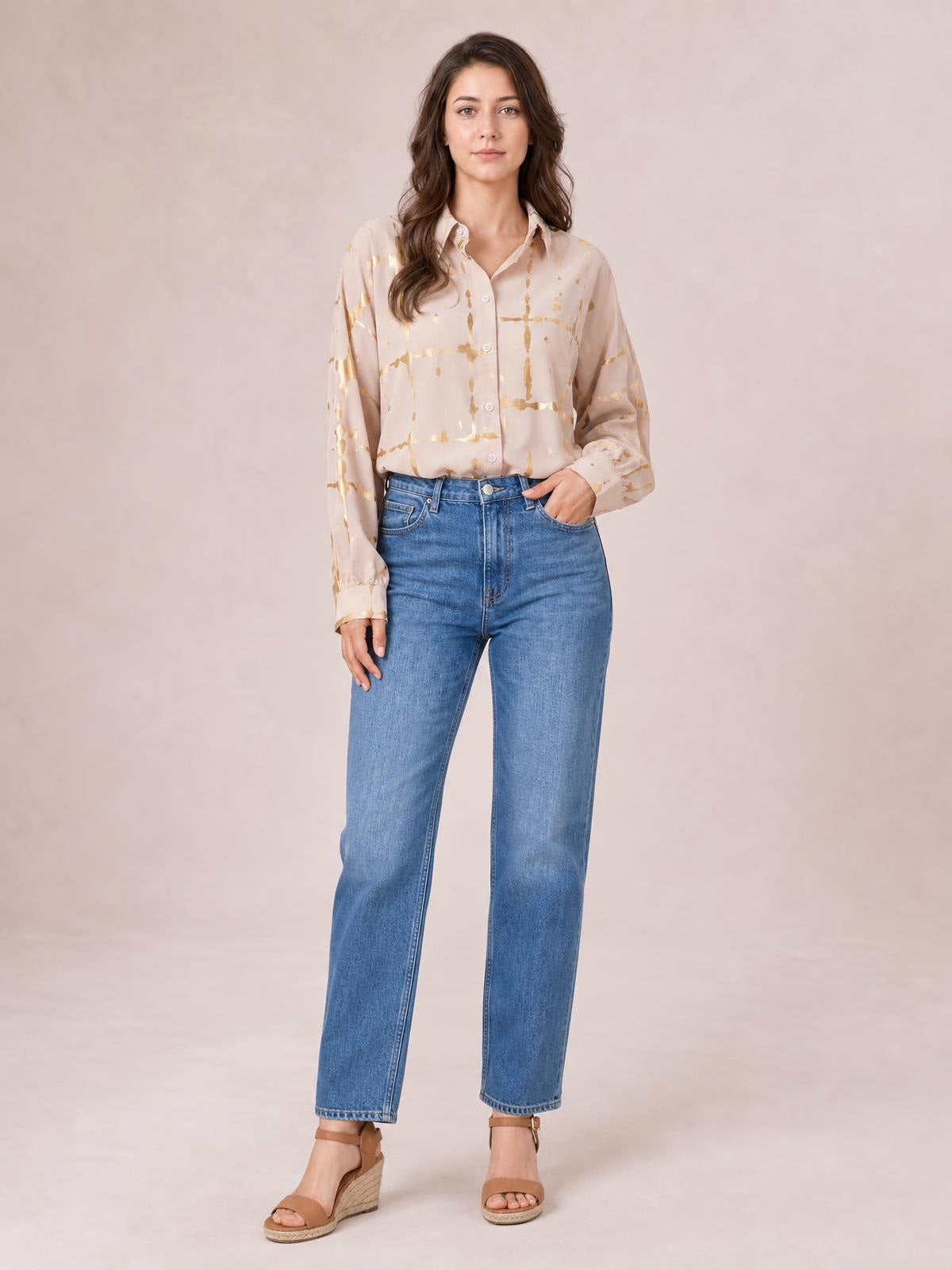 Model wearing a beige printed collared neck long sleeve shirt with a button front, relaxed silhouette, and gold abstract pattern, standing with one hand in her pocket.