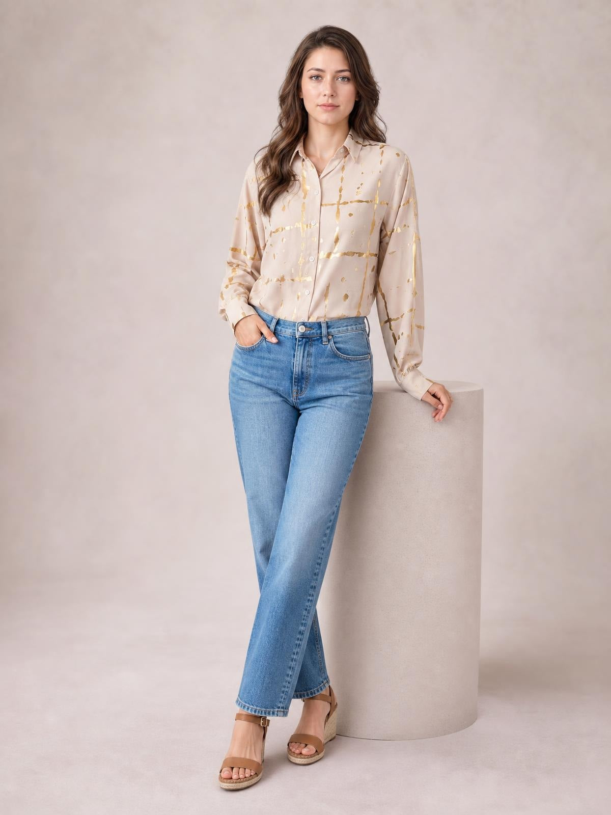 A model wears a blush printed collared neck long sleeve shirt with gold abstract lines, button-front construction, and a relaxed silhouette, leaning with one hand on a pedestal.