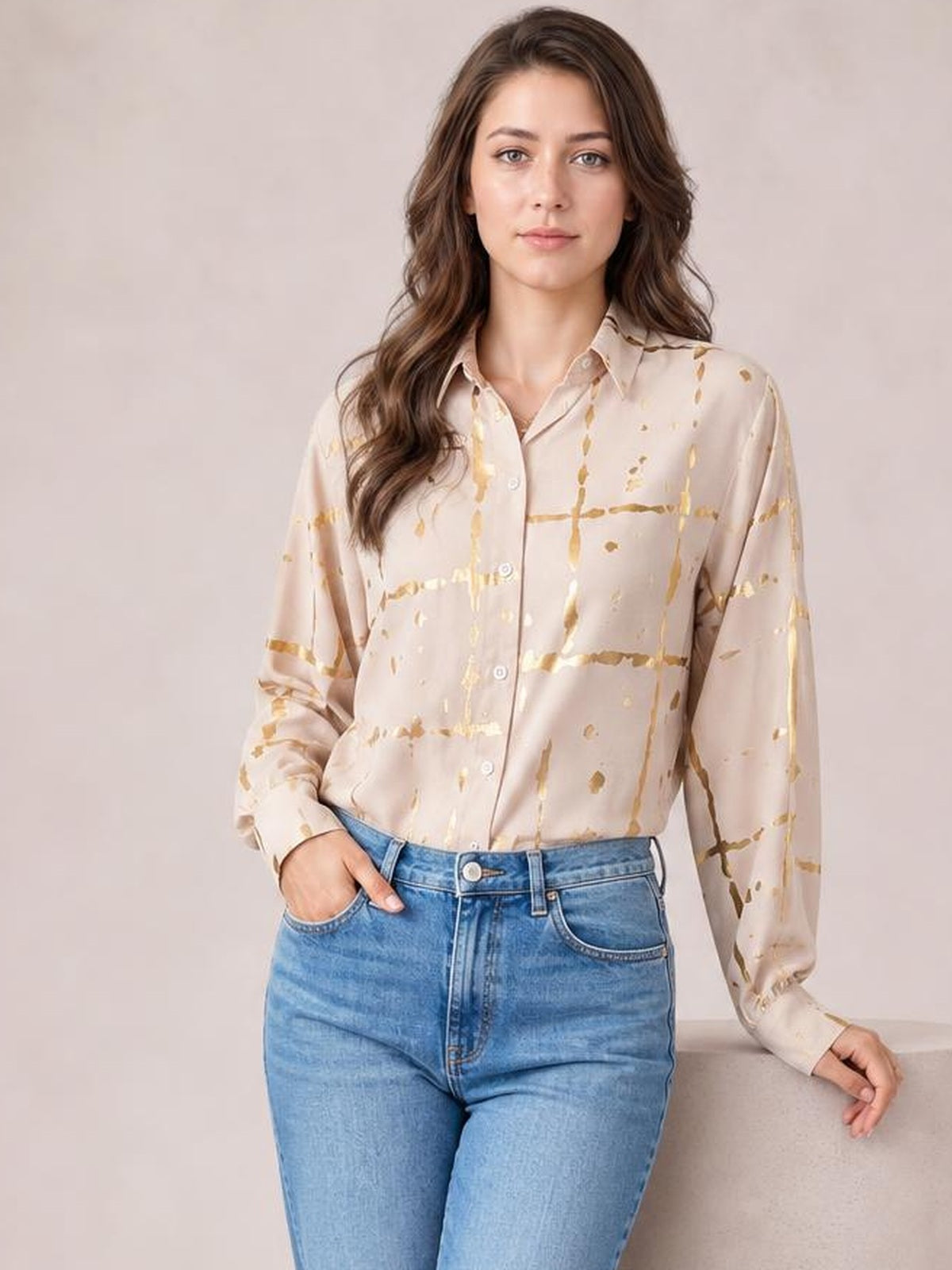 Model wearing a beige printed collared neck long sleeve shirt with button front and relaxed silhouette, paired with blue jeans and one hand in a pocket.