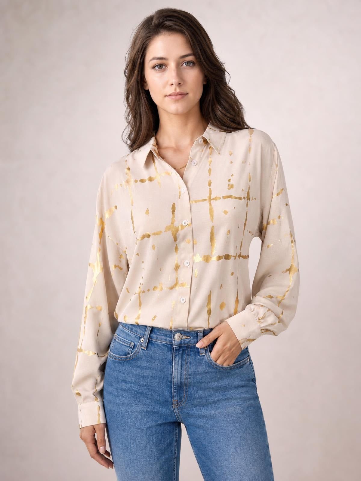 Model poses with one hand in her jeans pocket, wearing a cream printed collared neck long sleeve shirt with gold abstract grid detailing and a relaxed button-front silhouette.